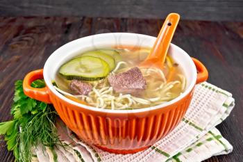 Soup with zucchini, beef, ham, lemon and noodles in a red bowl, parsley and dill on a towel on a wooden plank background