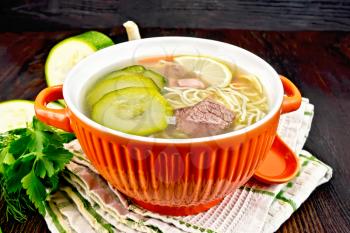Soup with zucchini, beef, ham, lemon and noodles in a bowl, parsley and dill on a kitchen towel on a wooden board background