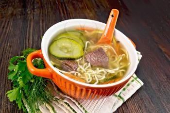 Soup with zucchini, beef, ham, lemon and noodles in a red bowl, parsley and dill on a kitchen towel on a wooden plank background