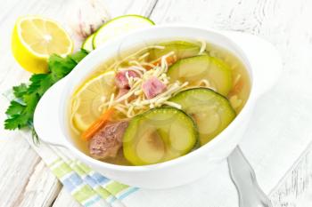 Soup with zucchini, beef, ham, lemon and noodles in a bowl on a towel, parsley and dill on a wooden plank background