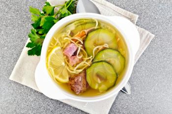 Soup with zucchini, beef, ham, lemon and noodles in a bowl on a napkin, parsley and dill on a granite table background on top