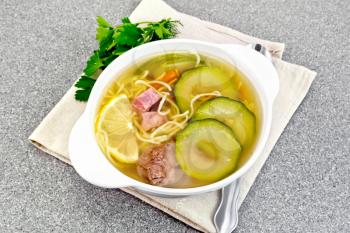 Soup with zucchini, beef, ham, lemon and noodles in a bowl on a napkin, parsley and dill on a gray granite table background