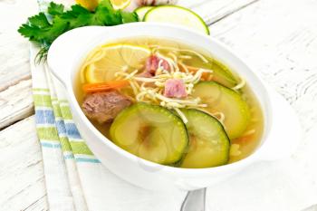 Soup with zucchini, beef, ham, lemon and noodles in a bowl on a napkin, parsley and dill on a light wooden board background