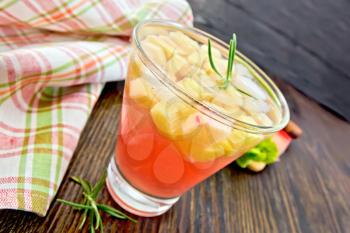 Lemonade with rhubarb and rosemary in a glass, stems and a leaf of a vegetable, checkered napkin on a wooden plank background