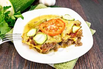 Casserole of minced meat, tomato and zucchini in a plate on a napkin, fork, garlic on a background of a dark wooden board