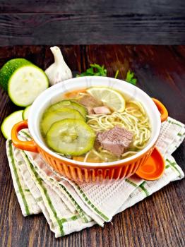 Soup with zucchini, beef, ham, lemon and noodles in a bowl, parsley and dill on a napkin on a wooden board background
