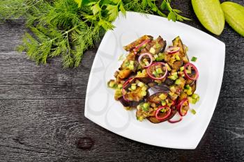 Salad of fried eggplant, fresh and pickled cucumber with red onion, seasoned with vegetable oil and spicy sauce in a plate, towel, fork and dill on a black wooden board background from above