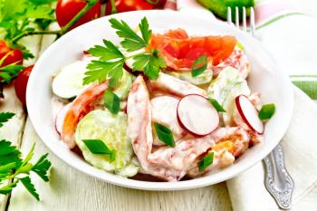 Salad of fresh tomatoes, cucumbers and radish with green onions and parsley,  flavored with mayonnaise and sour cream in a plate, napkin and fork on the background of a light wooden board