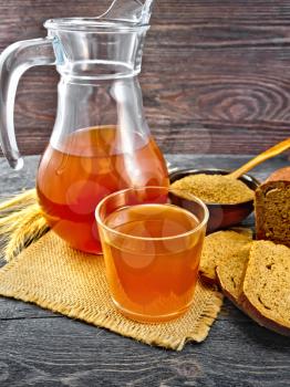 Kvass in glassful and glass jug on burlap, malt in a bowl, rye bread on a background of a dark wooden board