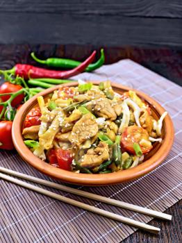 Thai noodles wok with chicken meat, tomatoes, soy sauce and green beans sprinkled with sesame seeds in a plate on a bamboo napkin with chopsticks on a wooden board background