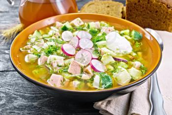 Cold soup okroshka from sausage, potato, egg, radish, cucumber, greens and kvass in a bowl, kitchen towel, bread and jug with drink on the background of wooden board