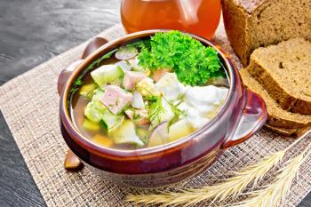 Cold soup okroshka from sausage, potatoes, eggs, radish, cucumber, greens and drink of kvass in a clay bowl, bread on brown wicker napkin on the background of wooden board