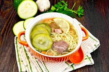 Soup with zucchini, beef, ham, lemon and noodles in a bowl, parsley and dill on a towel against a dark wooden board