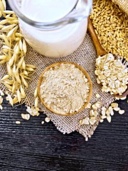 Flour oat in bowl, milk in jug, oatmeal in spoon on burlap, grain in bag, oaten stalks against a dark wooden board on top