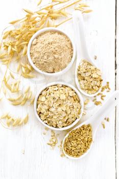 Oat flakes and flour in bowls, grain in a spoon, ripe stalks of oaten on a wooden board background from above