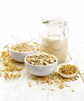 Oat flakes and flour in bowls, grain in a spoon, oatmeal milk in a glass jug and oaten stalks on a wooden board background