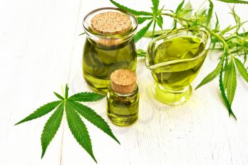 Hemp oil in two glass jars and sauceboat, leaves and stalks of cannabis on the background of white wooden boards