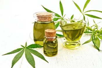 Hemp oil in two glass jars and sauceboat, leaves and stalks of cannabis on the background of light wooden boards
