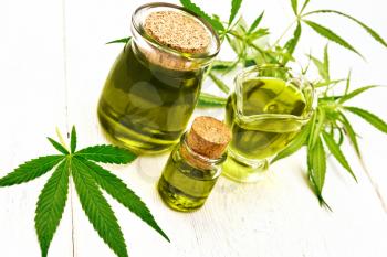 Hemp oil in two glass jars and sauceboat, leaves and stalks of cannabis on the background of wooden boards