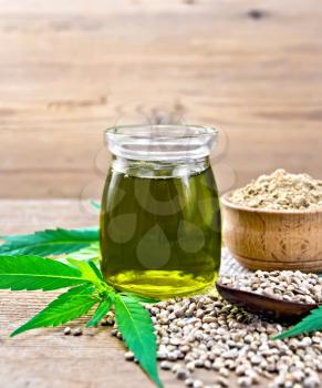 Hemp oil in a glass jar, grain in a spoon and flour in a bowl on sackcloth, cannabis leaves on the background of an old wooden board