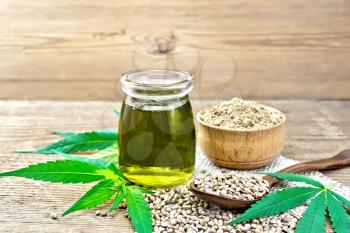 Hemp oil in a glass jar, grain in a spoon and flour in a bowl on a napkin of burlap, cannabis leaves on the background of an old wooden board