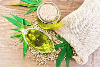 Hemp oil in a glass sauceboat and a jar with grain in a bag, leaves and stalks of cannabis on wooden board background from above