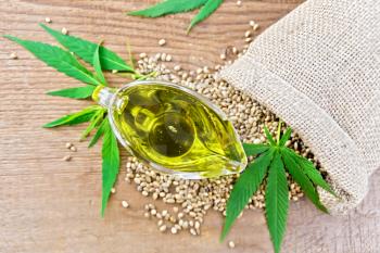 Hemp oil in a glass sauceboat with grain in a bag, leaves and stalks of cannabis against the background of an old wooden board from above