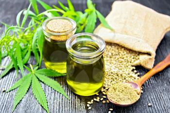 Hemp oil in two glass jars, grain in a bag and on the table, flour in a spoon, leaves and stalks of cannabis against the background of a dark wooden board