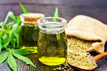 Hemp oil in two glass jars, grain in a bag and on the table, flour in a spoon, cannabis leaves against a dark wooden board