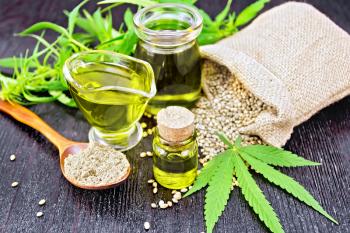 Hemp oil in two glass jars and sauceboat with grain in a sack, a spoon with flour, leaves and stalks of cannabis on a wooden board background