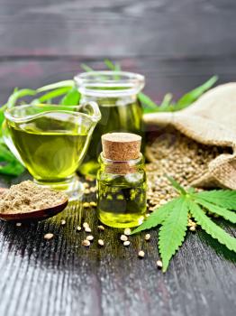 Hemp oil in two glass jars and sauceboat with grain in a sack, a spoon with flour, leaves and stalks of cannabis on a dark wooden board background