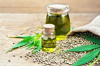 Hemp oil in two glass jars with grain in the sack, leaves and stalks of cannabis on the background of old wooden boards