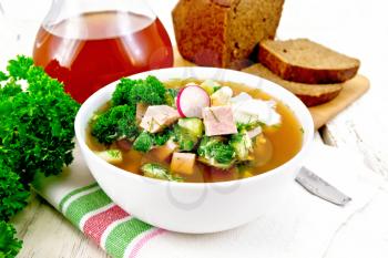Cold soup okroshka from sausage, potato, egg, radish, cucumber, greens and kvass in a white bowl on towel, bread and jug with drink on the background of light wooden board