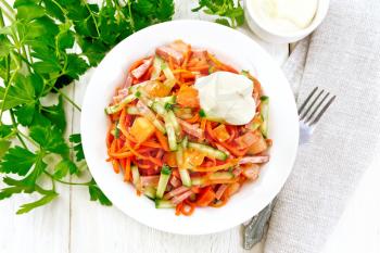 Salad from smoked sausage, spicy carrot, tomato, cucumber and spices with mayonnaise, napkin, fork and parsley on a light wooden board background from above