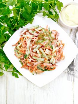 Salad from smoked sausage, spicy carrot, tomato, cucumber and spices, dressed with mayonnaise, napkin, fork and parsley on the background of a light wooden board from above
