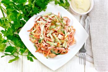 Salad from smoked sausage, spicy carrot, tomato, cucumber and spices, dressed with mayonnaise, napkin, fork and parsley on a wooden board background from above