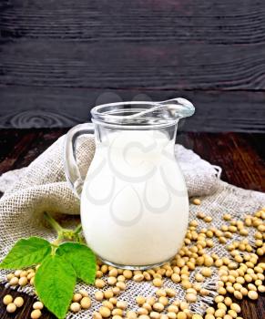 Soy milk in a jug, green leaf, soybeans on a napkin of burlap on a wooden board background