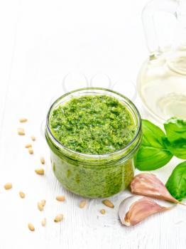 Pesto sauce in a glass jar, pine nuts, garlic, green basil and olive oil in a carafe on the background of light wooden board