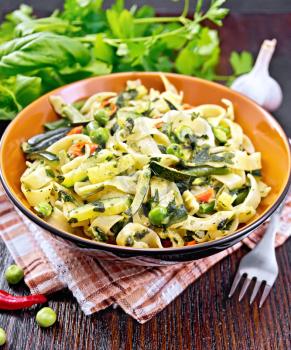 Tagliatelle pasta with zucchini, green peas, asparagus beans, hot pepper and spinach in a plate on napkin, garlic, fork and basil on wooden board background