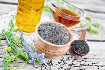 Flour of black caraway in a bowl, seeds in a spoon, oil in bottle and sauceboat on burlap napkin, sprigs of Nigella sativa with blue flowers and green leaves on background of wooden board