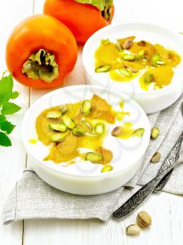 Dessert of yogurt, persimmon and honey with vanilla, cardamom and pistachios in two bowls on a towel, mint and spoon on wooden board background