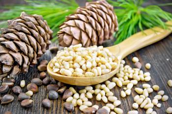 Peeled kernels of cedar nuts in a spoon, two pine cones, green branches and nuts in the shell on a wooden board background