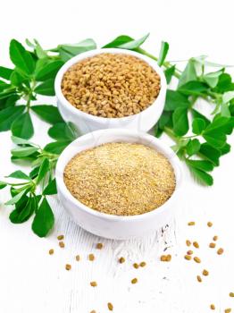 Fenugreek seeds and ground spice in two bowls and on a table with leaves on wooden board background