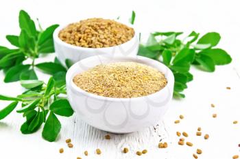 Fenugreek seeds and ground spice in two bowls and on a table with green leaves on wooden board background