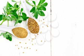 Fenugreek seeds and ground spice in two spoons and on a table with green leaves on a wooden board background from above