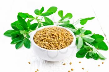 Fenugreek seeds in a bowl and on a table, green seasoning leaves on white wooden board background