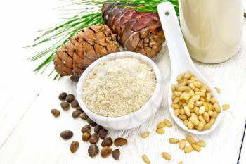 Cedar flour in a bowl, nuts and cones, a spoon with peeled nuts, pine branch and cedar milk in a bottle on wooden board background