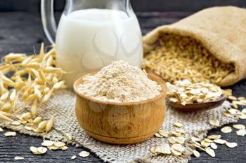 Flour oat in a bowl, milk in a jug, oatmeal in a spoon on burlap, grain in bag, oaten stalks on wooden board background
