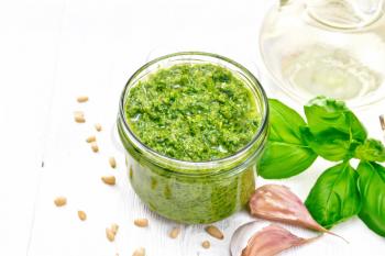 Pesto sauce in a glass jar, pine nuts, garlic, green basil and olive oil in a carafe on wooden board background