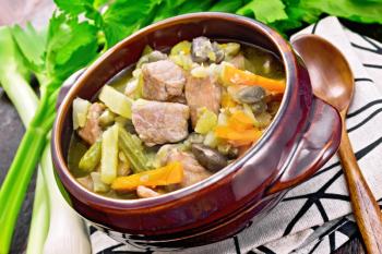Eintopf soup of pork, celery, beans, carrots and potatoes with leek in a clay bowl on a towel on background of dark wooden board